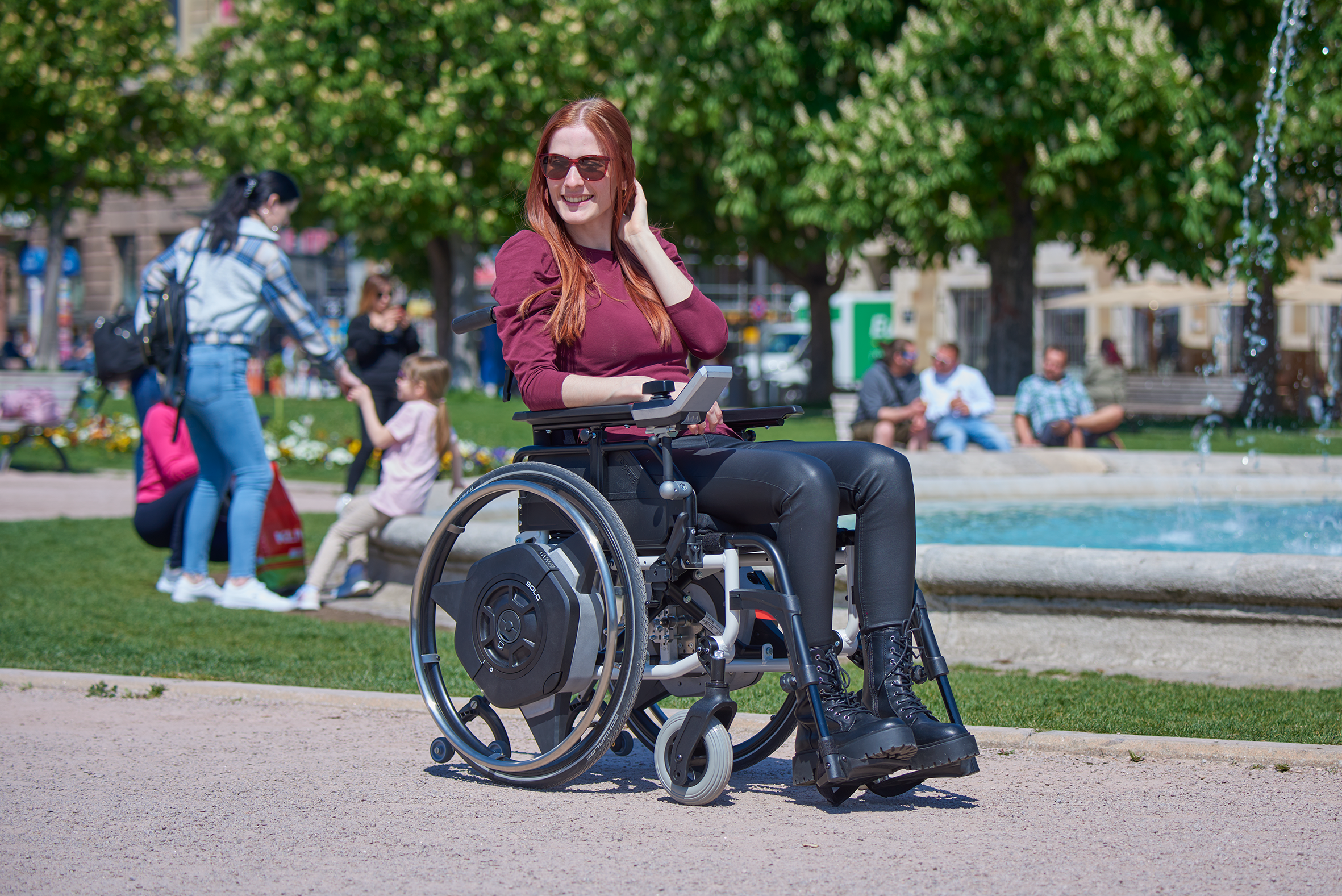 Das Bild zeigt eine junge Frau, die mit ihrem manuellen Rollstuhl inklusive dem neuesten Zusatzantrieb fürü Rollstühle SOLO², in einem belebten Park unterwegs ist.